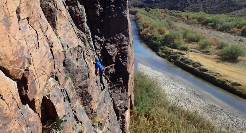 A person wearing safety gear climbs up a rock face high above a river below. 